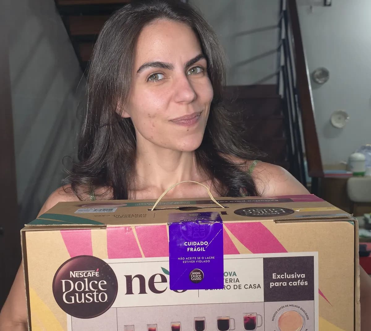 Cecília segurando a caixa lacrada da máquina NESCAFÉ Dolce Gusto neo recém-entregue, com o selo "não aceite se o lacre estiver violado" visível